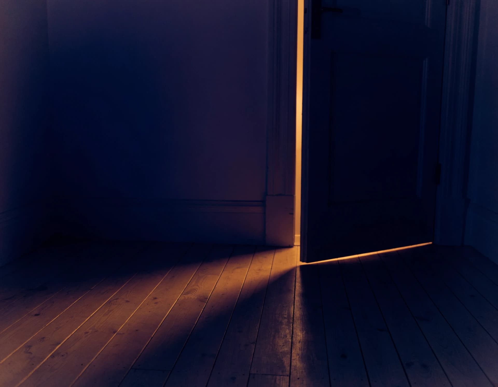 A single thin blade of warm light falling across the bare floor of a dark, empty room through the gap of a nearly-closed door.