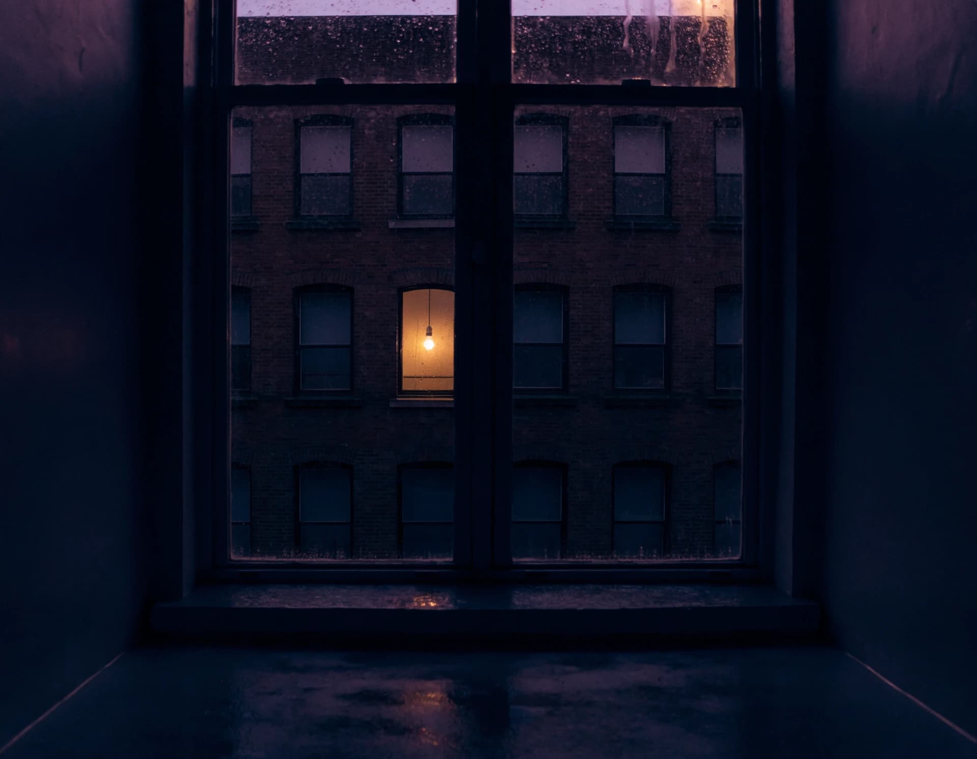 Seen from a dark empty room, a single warm-lit window in the dark building across the way beneath a violet pre-dawn sky.